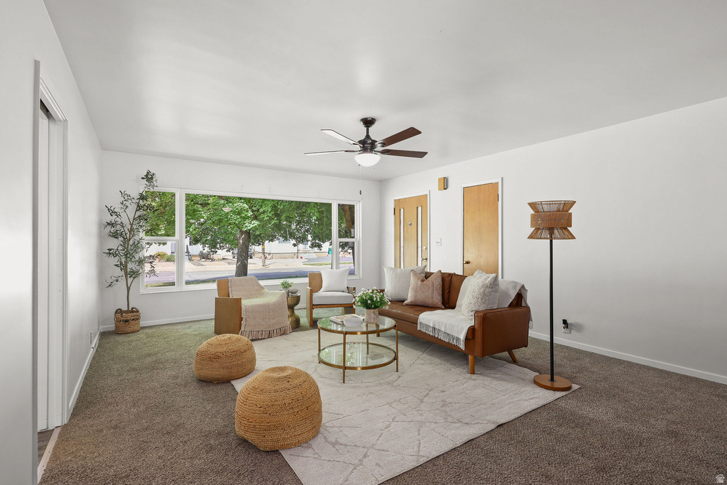 Bright living area featuring a large picture window, ceiling fan with light fixture, light brown carpeting, and white walls
