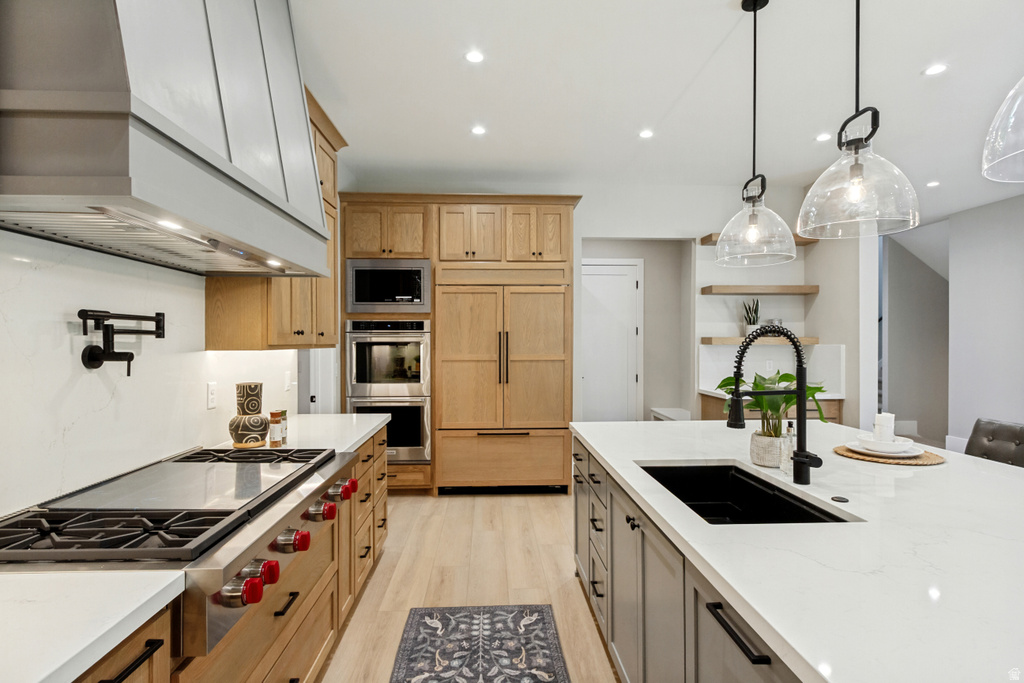 Gourmet kitchen featuring a substantial island with an undermount sink, pendant lighting, professional-grade range with a custom hood, built-in double ovens, and light wood-finish cabinetry