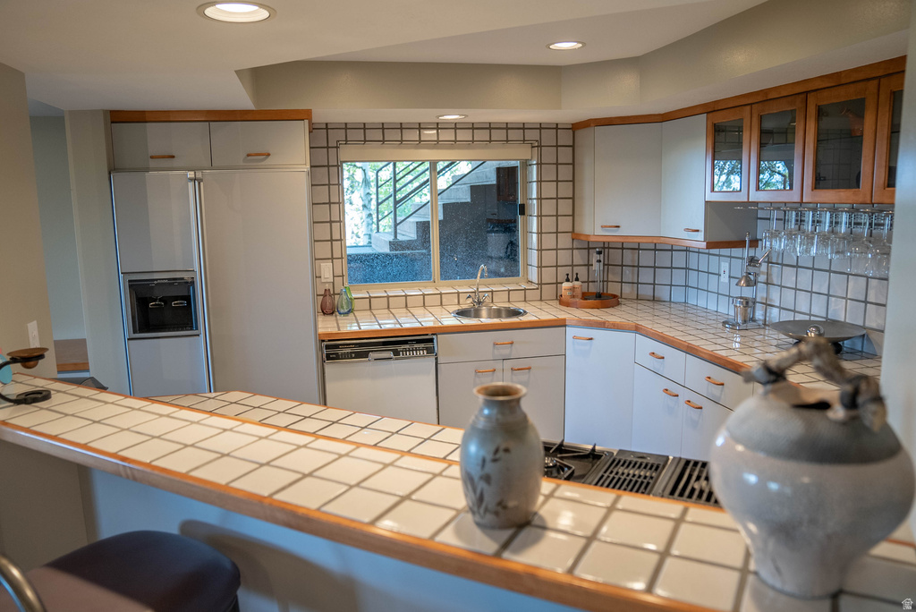 Kitchen featuring white tiled countertops and backsplash, white cabinetry, integrated refrigerator with water dispenser, stainless steel sink, and wood-framed upper cabinets with glass inserts