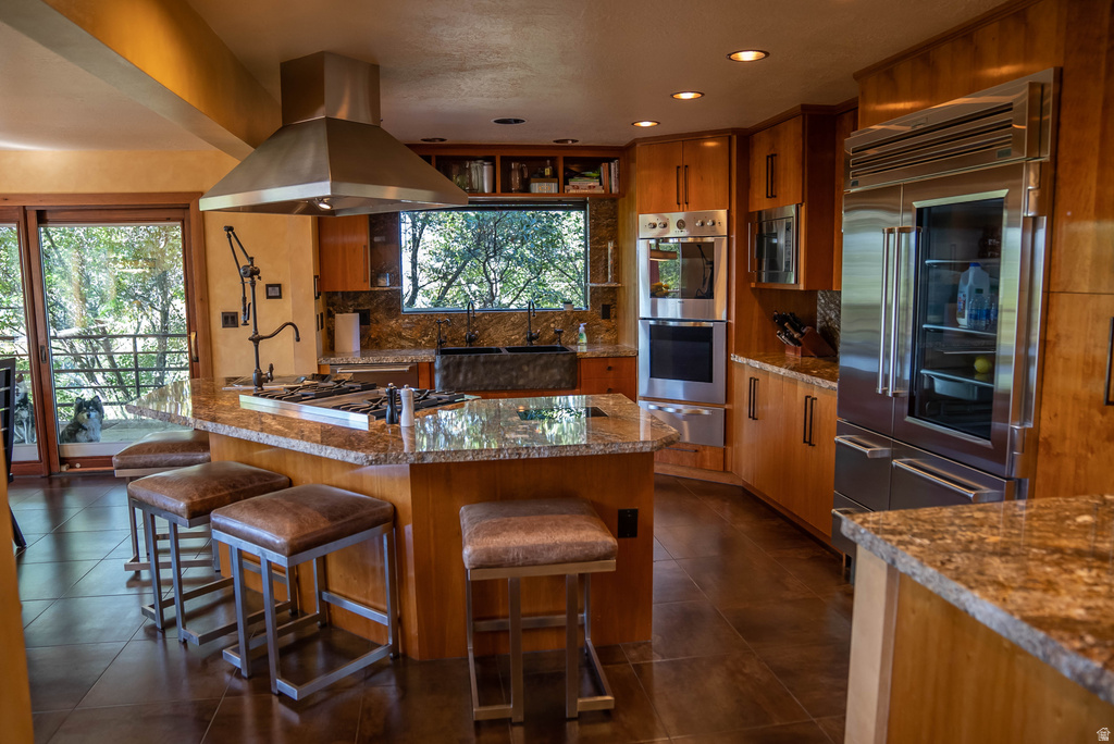 Gourmet kitchen featuring a large island with granite countertops, integrated gas cooktop, stainless steel range hood, built-in double ovens, and a paneled refrigerator with glass-front compartment