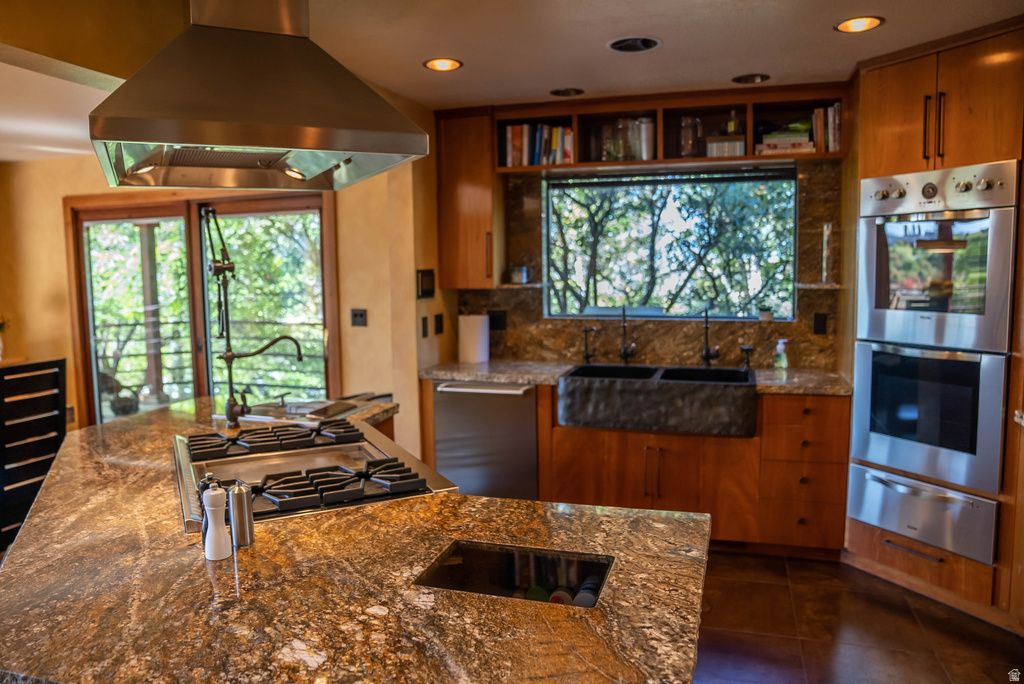 Gourmet kitchen featuring a central island with an integrated cooktop and stainless steel hood, rich wood cabinetry, granite countertops, and built-in double ovens