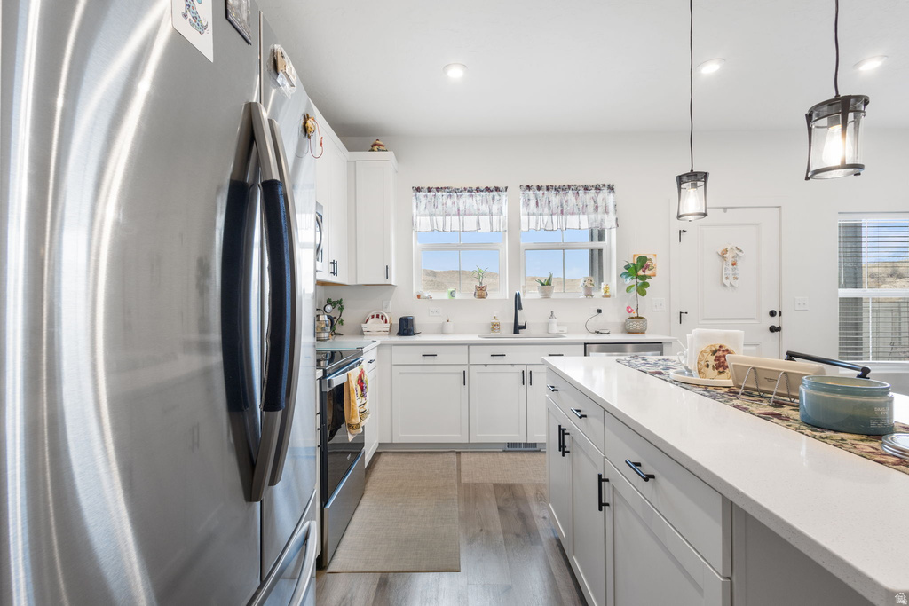 Kitchen with stainless steel appliances, white cabinets, decorative light fixtures, light wood-type flooring, and light stone counters