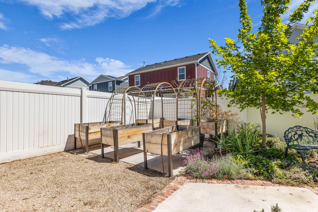 Fenced backyard featuring multiple raised garden beds with metal arch trellises, a young tree, and a concrete patio with a brick border