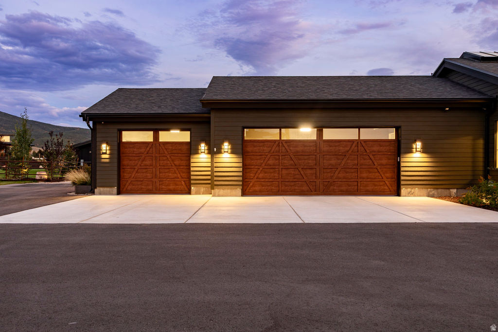 Two-car garage featuring rich wood-finish doors with upper window panels, dark siding, multiple exterior wall sconces, and a concrete driveway