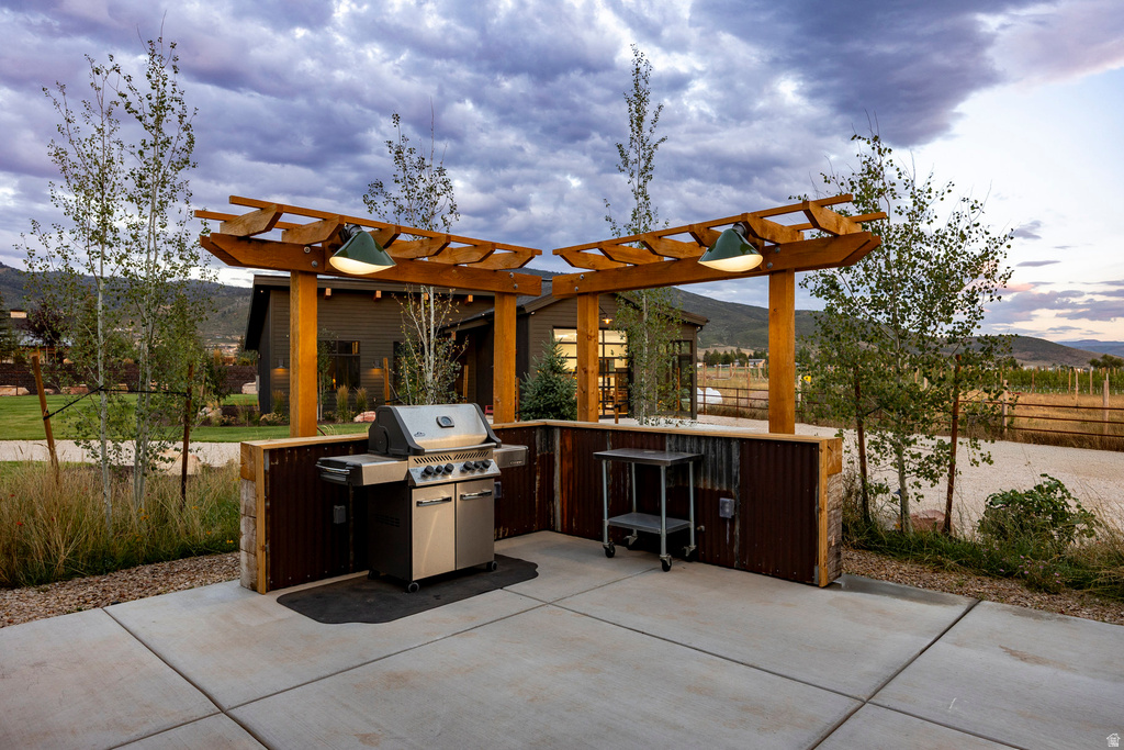 Outdoor kitchen area featuring a built-in stainless steel grill, a concrete patio, a wood pergola with integrated lighting, and a stone and corrugated metal half-wall