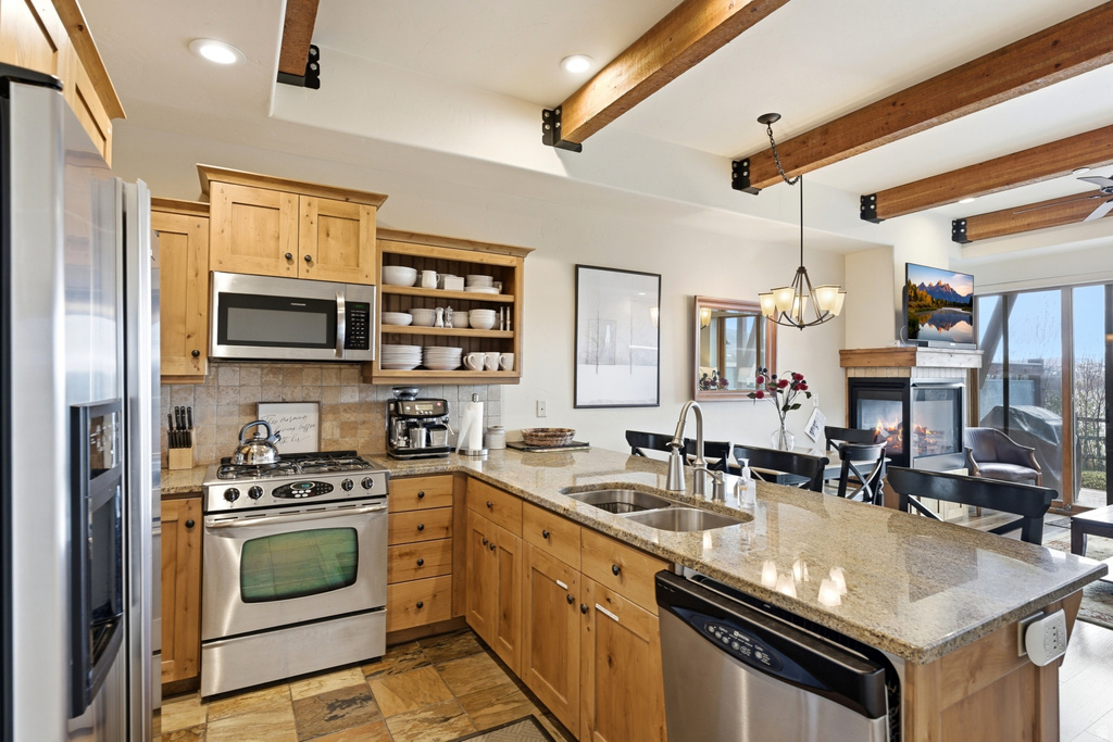 Kitchen featuring light wood cabinetry, stainless steel appliances, stone tile flooring, and a stone countertop with a double basin sink