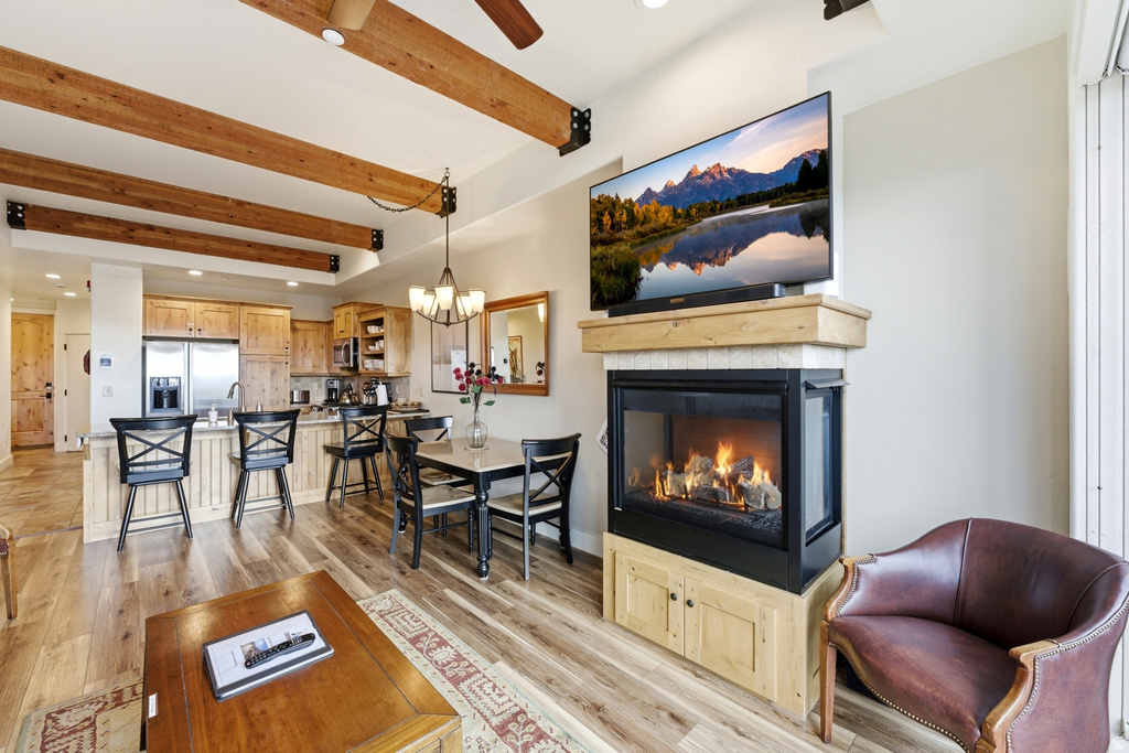 Open-concept living area featuring exposed wood ceiling beams, wood-finish flooring, a three-sided fireplace with natural wood base cabinetry, and a full kitchen with stainless steel appliances
