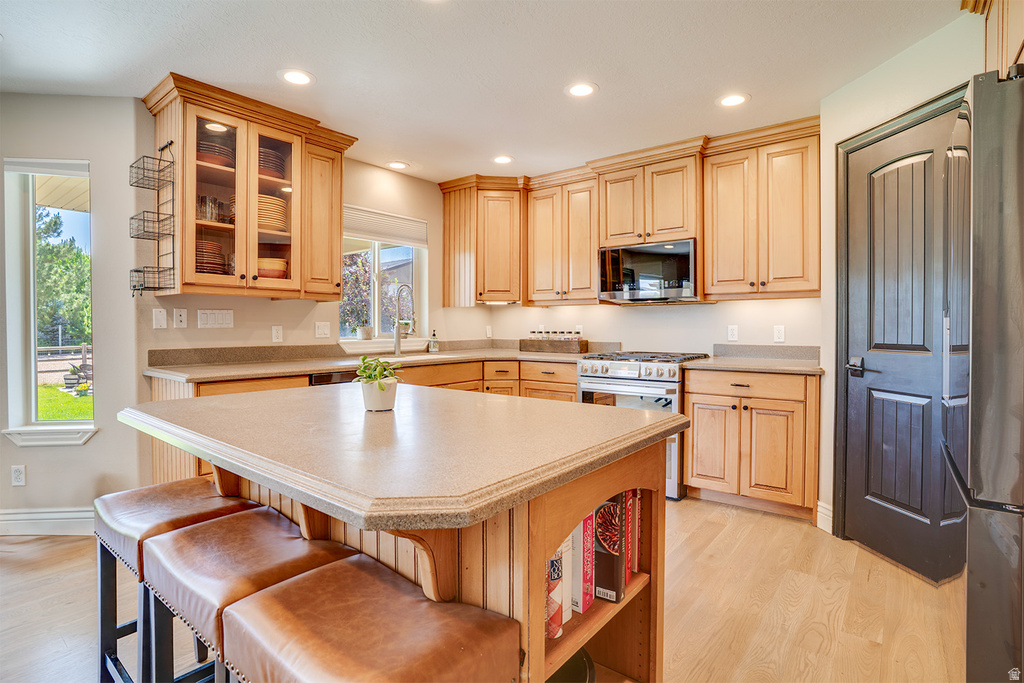 Spacious kitchen featuring light wood cabinetry, a central island with built-in shelving, stainless steel appliances including a gas range and microwave, and light-toned countertops