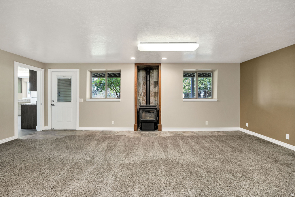Spacious room featuring neutral paint, soft grey carpeting, a corner wood-burning stove, two windows, and a doorway leading to an additional space with wood-finish cabinetry