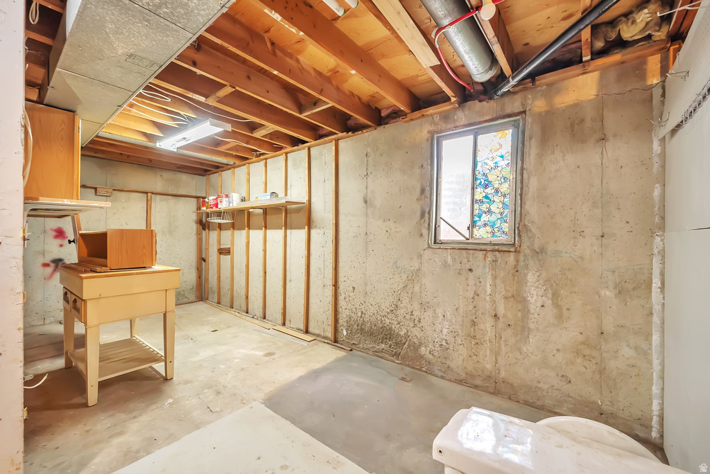 Unfinished basement space featuring exposed wooden ceiling joists, concrete flooring, a single window, and visible plumbing and ductwork