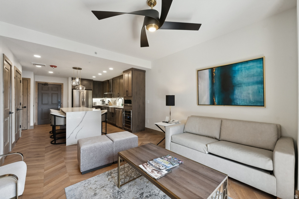 Open-concept living space featuring wood-finish herringbone flooring, a modern kitchen with dark wood cabinetry, stainless steel appliances, a waterfall island with stone countertop, and a contemporary ceiling fan with integrated lighting