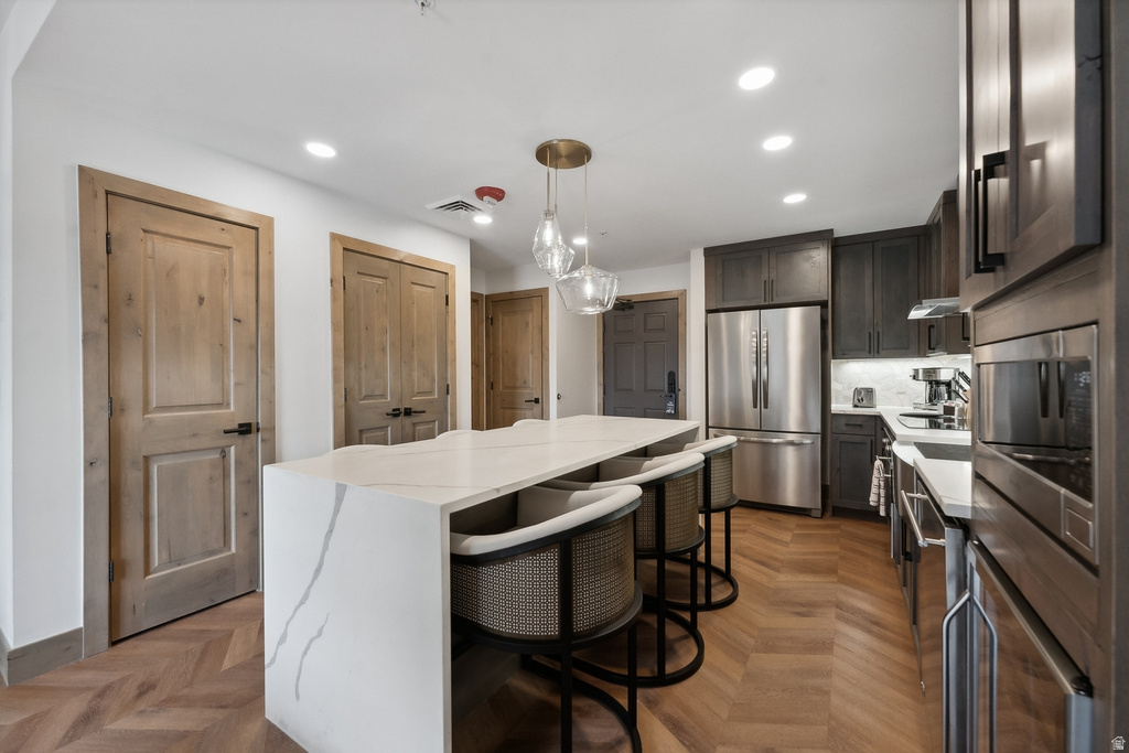 Kitchen featuring a waterfall-edge island with seating, integrated stainless steel appliances, dark wood-finish cabinetry, and chevron-patterned wood-finish flooring
