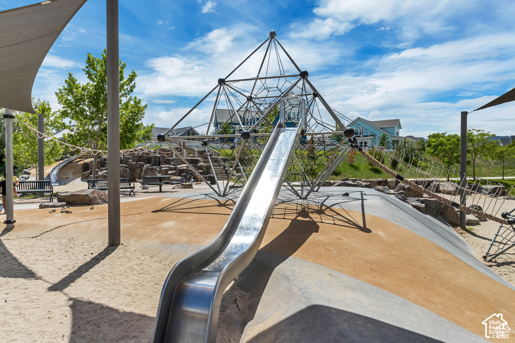 Contemporary playground featuring a large metal slide, geometric climbing structure, rope bridge, shade sails, and sand ground cover