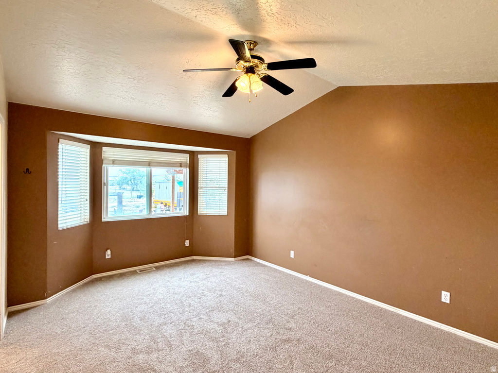 Spacious interior room featuring a large bay window with horizontal blinds, sloped ceiling, ceiling fan with integrated lighting, and neutral carpet flooring