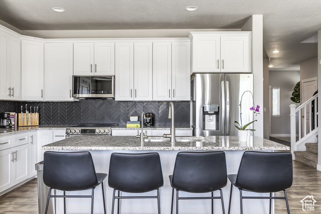 Modern kitchen featuring white cabinetry, stainless steel appliances, a dark herringbone tile backsplash, and a granite island with an integrated sink