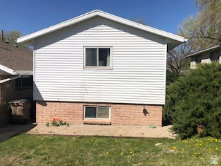 White siding exterior with a pitched roof, featuring brick foundation accents, a ground-level window, and a mature evergreen shrub
