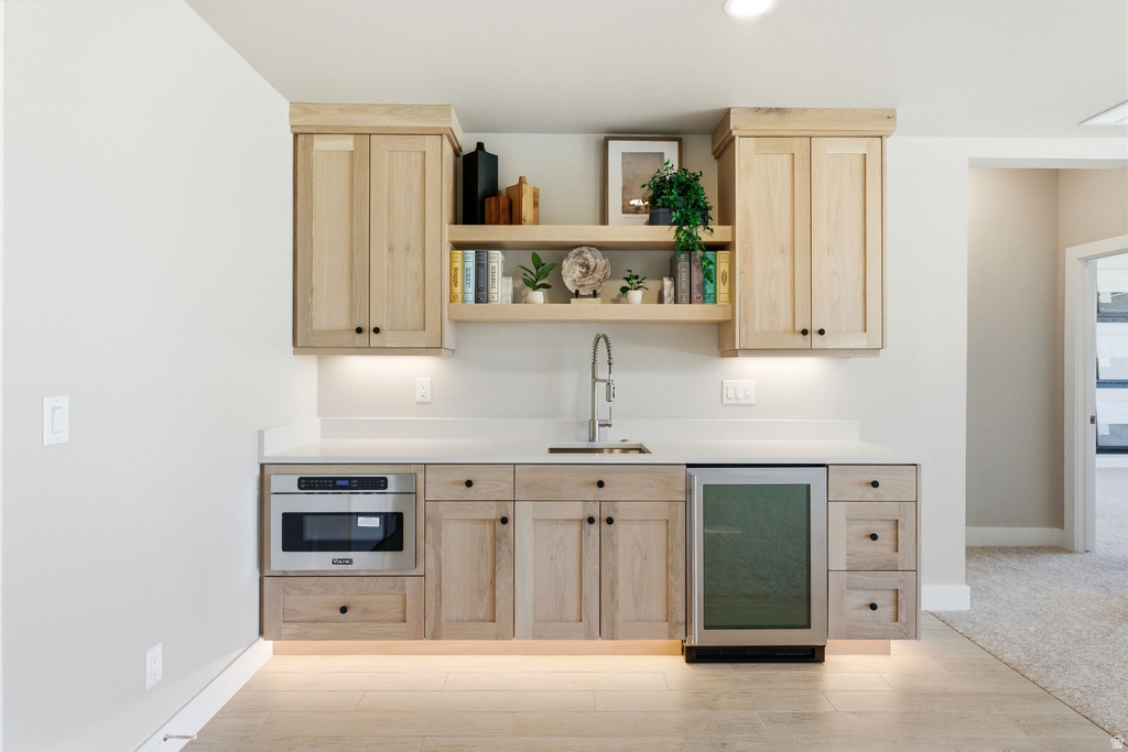 Built-in wet bar featuring light wood-finish cabinetry, under-cabinet lighting, a stainless steel microwave, a beverage cooler, and a single basin sink with a gooseneck faucet