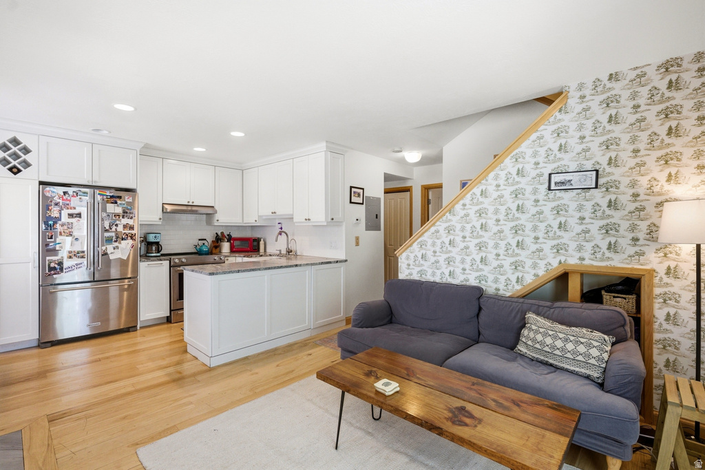 Open-concept living area featuring light wood-finish flooring, white shaker-style cabinetry, stainless steel appliances, a kitchen island with a light-colored countertop, and a wooden staircase with a half-wall railing