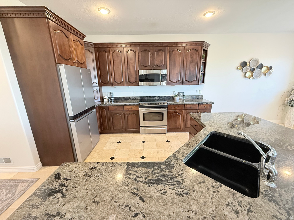 Kitchen featuring a large central island with a double basin sink, dark wood cabinetry with crown molding, stainless steel appliances, and light-toned tile flooring with dark accent tiles
