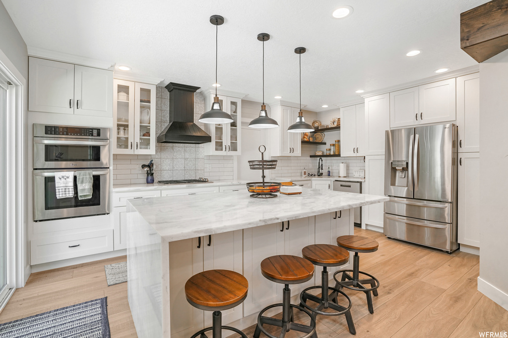 Bright kitchen featuring a waterfall island with stone countertop, stainless steel appliances including a double oven and refrigerator, white cabinetry, and wood-finish flooring