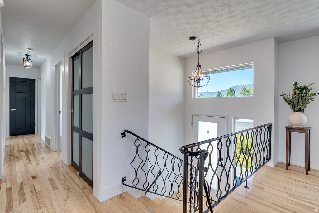 Upper level hallway featuring light wood-finish flooring, a contemporary chandelier, a large window showcasing distant mountains and trees, a decorative wrought-iron staircase railing, and a modern frosted glass door