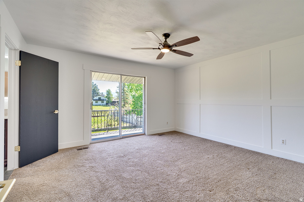Carpeted room featuring a ceiling fan, a white paneled accent wall, and a sliding glass door opening to an outdoor balcony with an iron railing