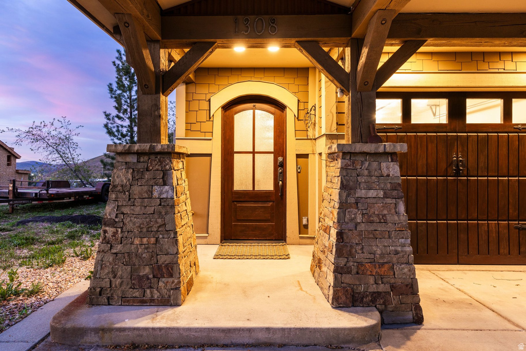 Inviting front entryway featuring stone-clad columns, exposed wood beam accents, a paneled arch-top entry door with glass inserts, and an integrated garage door with window panels