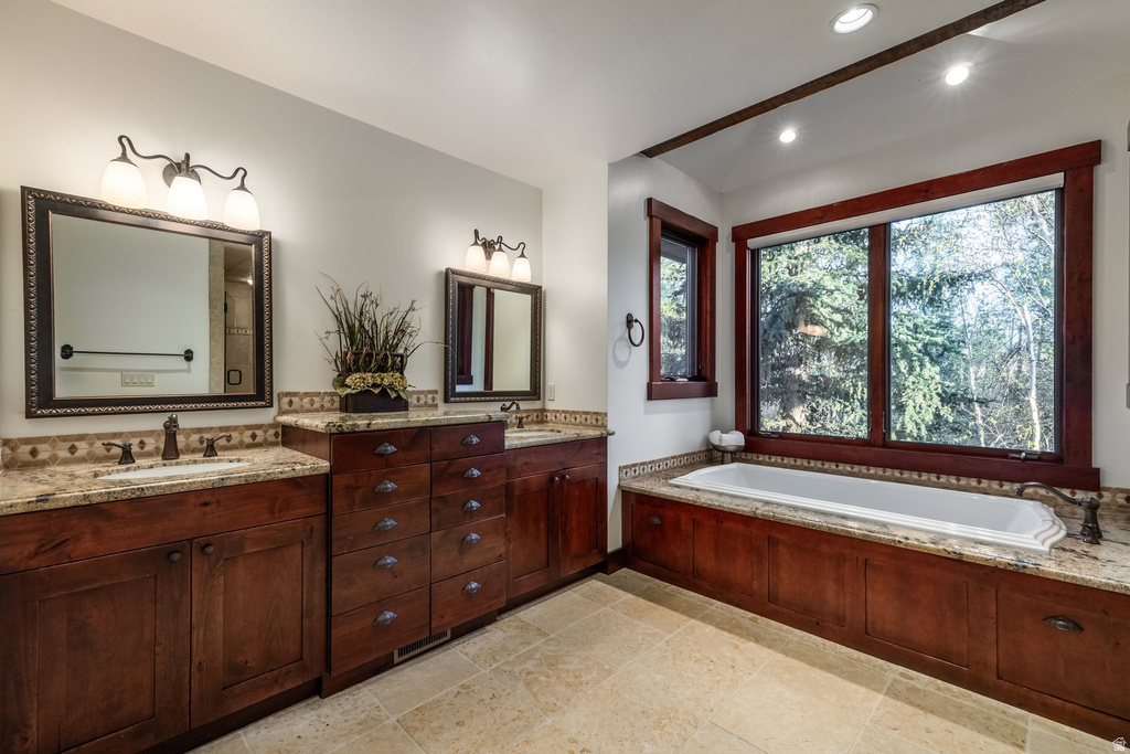 Spacious bathroom featuring a dual vanity with granite countertops, rich wood cabinetry, a built-in soaking tub, and large windows with wood trim