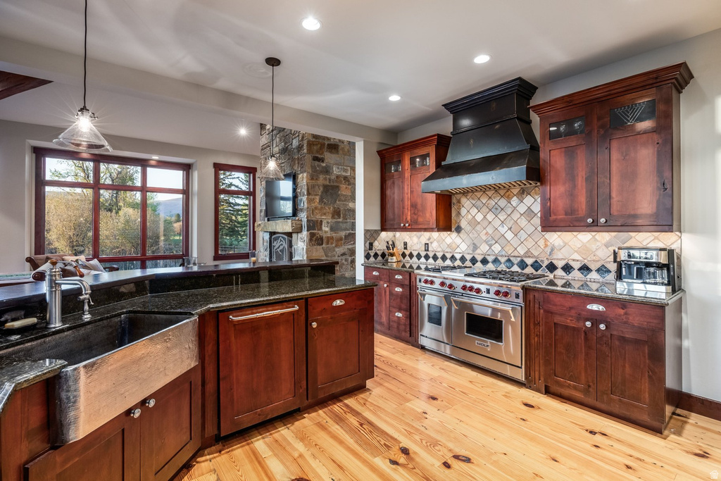Kitchen featuring a large farm sink, dark wood cabinetry, a professional-grade range with a custom hood, stone tile backsplash, and dark countertops