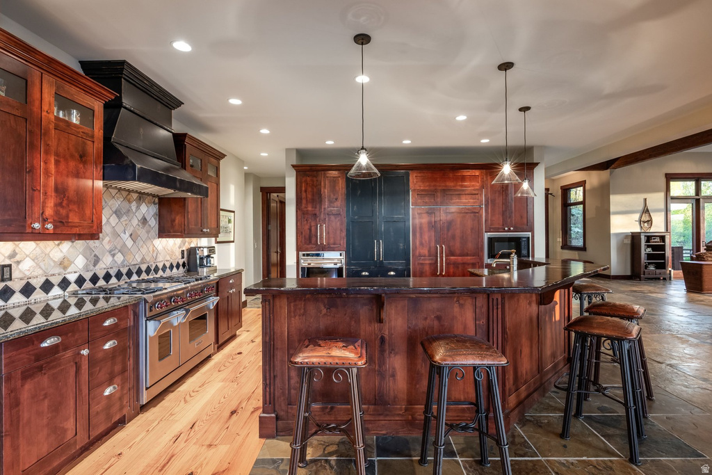 Gourmet kitchen with custom wood cabinetry, a large center island, professional-grade stainless steel range with custom hood, stone tile backsplash with decorative accents, and recessed and pendant lighting