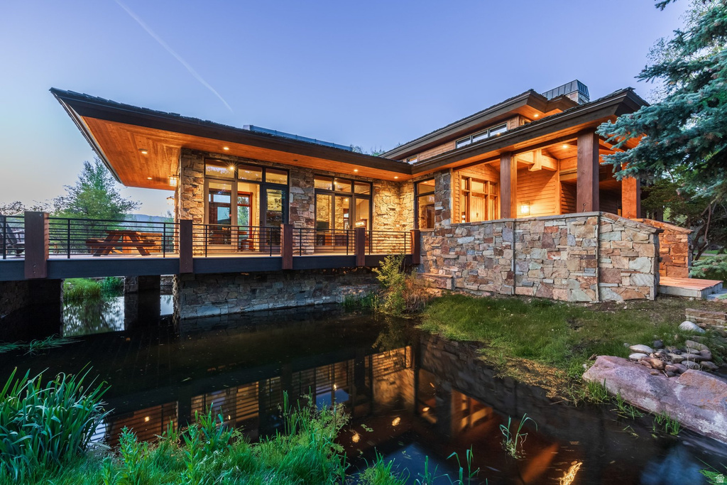 Contemporary exterior featuring stone cladding, a prominent deck over a water feature, extensive glass doors and windows, natural wood soffits, and a covered patio area