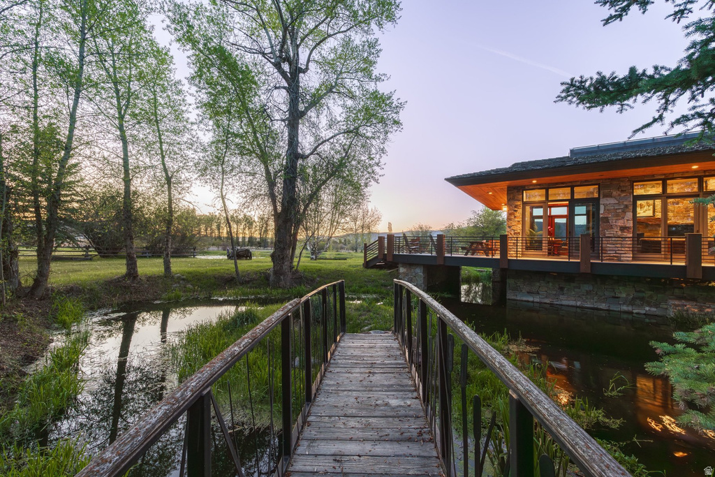 Wood-finish footbridge spanning a tranquil water feature, leading to a modern stone and wood-clad residence