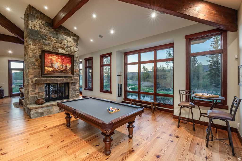 Spacious room featuring a prominent stone fireplace, wood-finish flooring, exposed ceiling beams, and large windows with dark wood trim