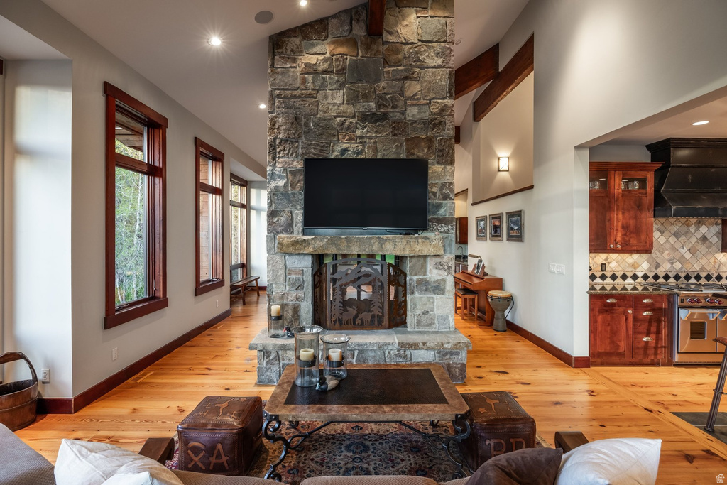 Central stone fireplace with a substantial hearth, wood-finish flooring, recessed lighting, exposed ceiling beams, and a kitchen featuring a tile backsplash and stainless steel appliances