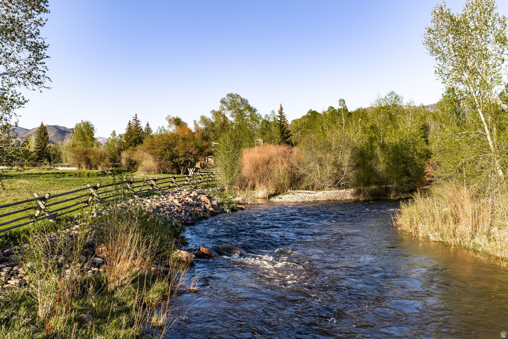 Riverfront property featuring a split-rail fence, a grassy expanse, mature trees, and mountainous terrain in the background