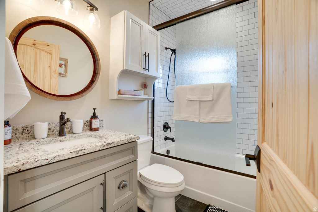 Bathroom featuring a vanity with a light gray cabinet and a speckled countertop, an oval mirror with a wooden frame, a white subway tile shower surround, a tub with a sliding frosted glass door, and a wooden door with dark hardware