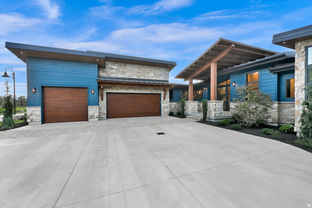 Modern exterior featuring blue horizontal siding, stone accents, two wood-finish garage doors, a covered entry with exposed wood beams, and a concrete driveway