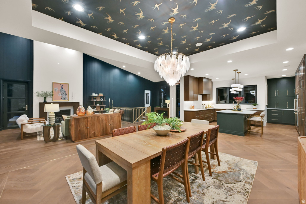 Open-concept living space featuring a distinct dining area with a feather-motif chandelier, a living room with a fireplace, and a kitchen with dark cabinetry