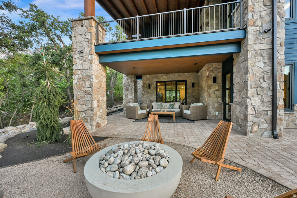 Ground-level patio featuring a fire pit, surrounding gravel and pavers, a covered outdoor seating area with built-in lighting, and stone columns supporting an upper-level balcony with metal railings