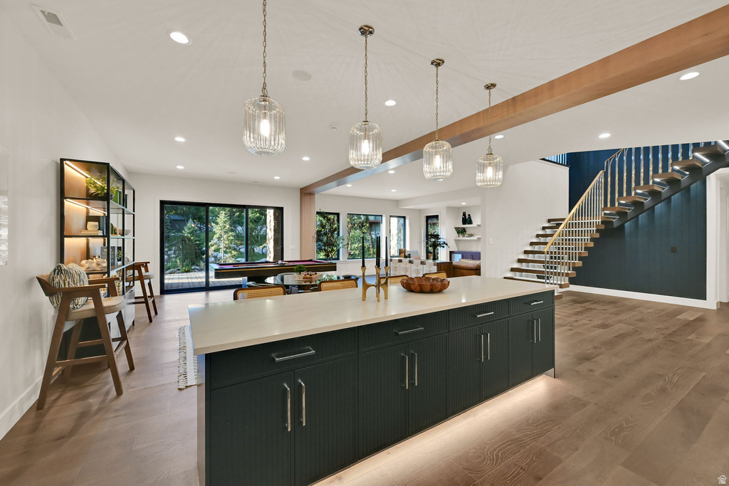 Spacious open-concept interior featuring a substantial island with dark cabinetry and a light countertop, wood-finish flooring, recessed lighting, and a prominent staircase with white railings