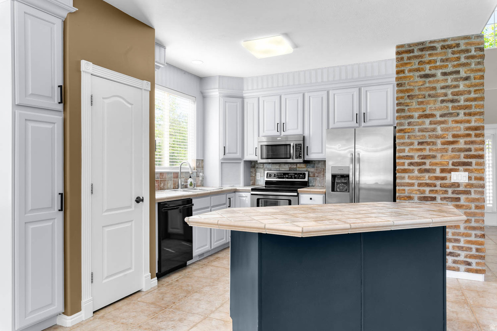 Kitchen featuring a tiled island, white cabinetry, stainless steel appliances, and a brick-finish accent wall
