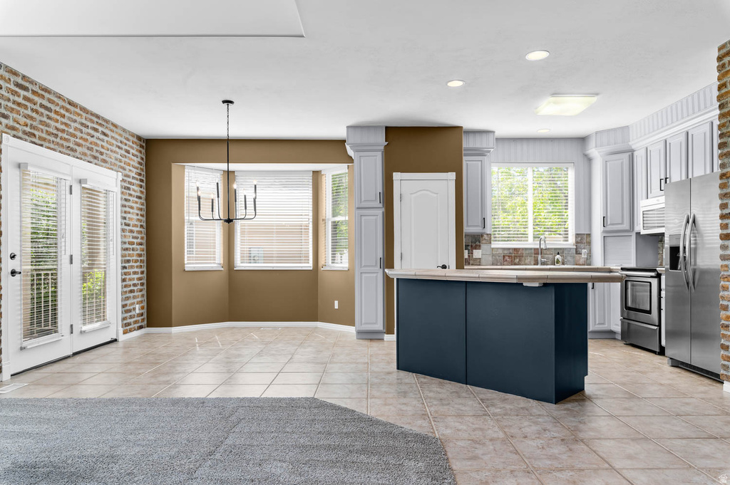 Open-concept living space featuring a central kitchen island with seating, a bay window with natural light, tiled flooring, and a brick accent wall with double French doors