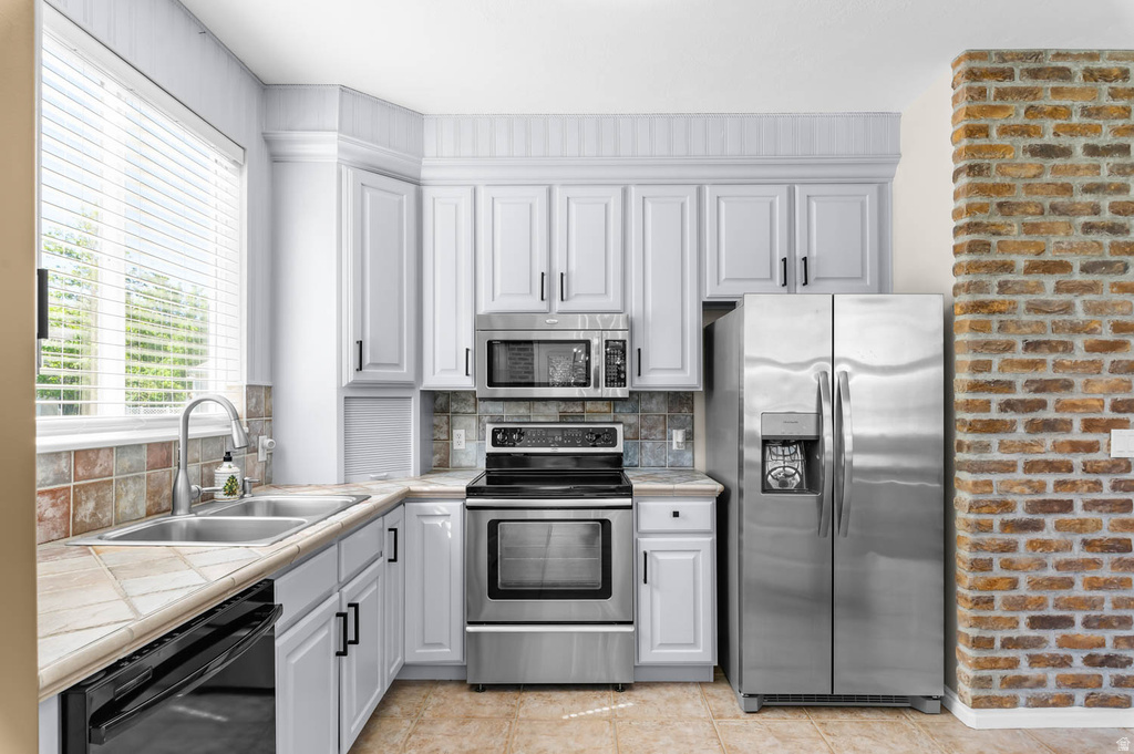 Kitchen featuring white cabinetry with dark hardware, stainless steel appliances, a double basin sink, tiled countertops, and a brick accent wall