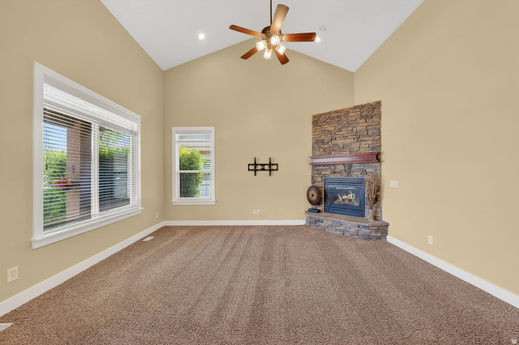 Spacious living area featuring a vaulted ceiling with a ceiling fan, a stone-clad fireplace with a wood mantel, ample natural light through multiple windows, and plush carpeting