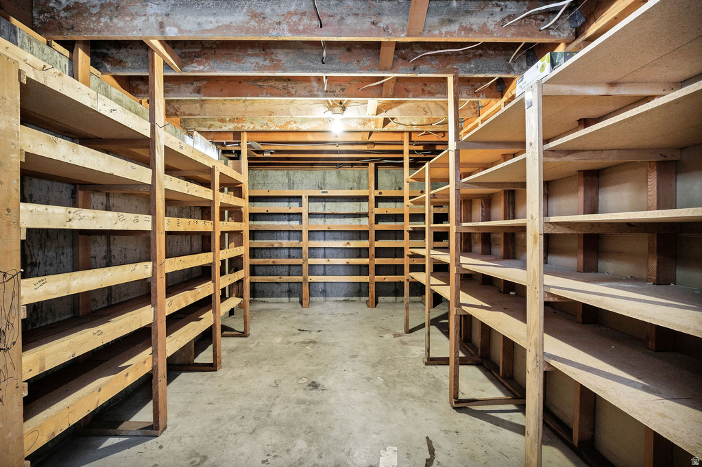 Basement storage area featuring extensive built-in shelving, exposed wood beam ceiling, concrete flooring, and a single ceiling-mounted light fixture