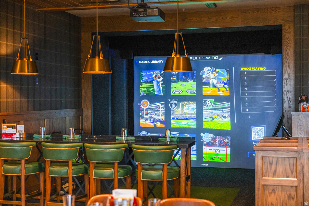 Indoor entertainment space featuring a large projection screen, dark paneled walls, wood trim, a bar with a dark countertop, and green upholstered bar stools