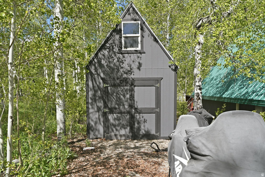 Gable-roof storage shed with dark gray siding, a white-framed window, and double-entry doors surrounded by mature trees