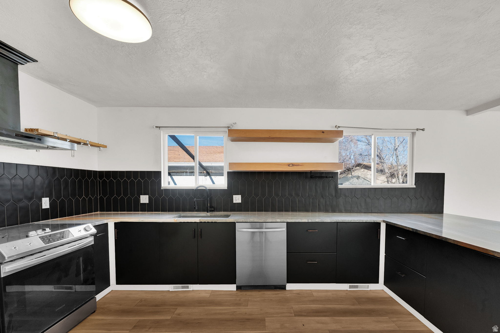 Modern kitchen featuring black cabinetry, a dark geometric tile backsplash, wood-finish flooring, and stainless steel appliances