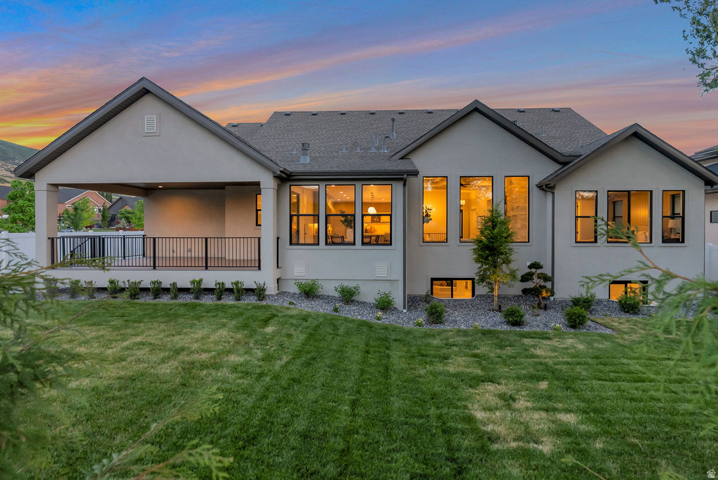 Contemporary stucco exterior featuring a covered patio with dark railings, a spacious lawn, multiple large windows, and lower-level windows