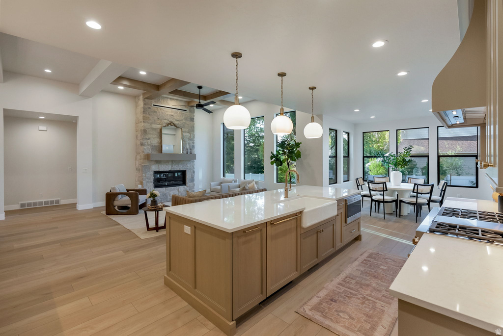 Open-concept living space featuring wood-finish flooring, a stone-faced fireplace, a coffered ceiling, a kitchen island with an integrated farmhouse sink, and a dining area with multiple large windows